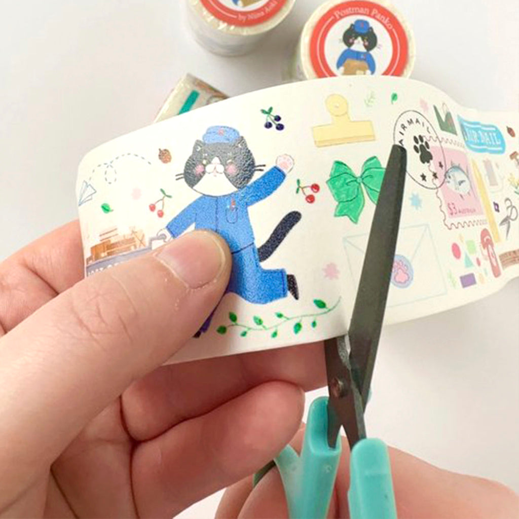 A hand holds a roll of decorative washi tape with a cartoon cat in a blue outfit and small colourful illustrations, while teal-handled scissors cut the tape. Other rolls of tape are blurred in the background.