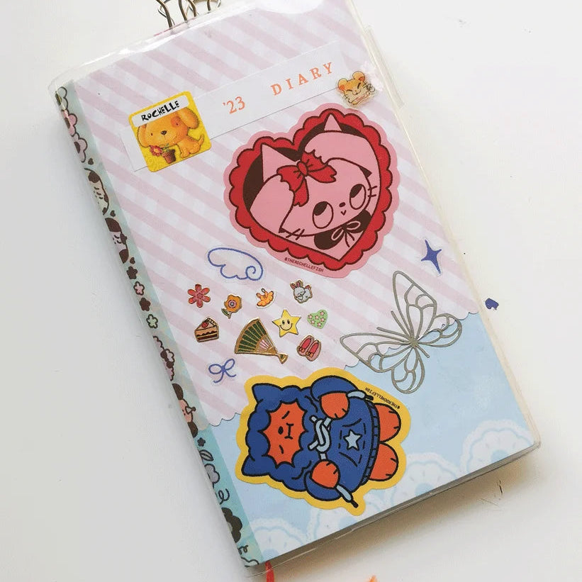 A diary with a pastel pink and light blue striped cover decorated with cute cartoon stickers, including a red cat face, a cat in blue outfit, and a butterfly. The notebook is sealed in a clear plastic sleeve with a metal binder clip at the top and an orange bookmark ribbon at the bottom.