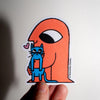 A hand holds a die-cut sticker featuring an orange figure with one large eye and a blue cat character, against a plain light background.