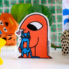 A die-cut sticker featuring an orange figure holding a blue cat, displayed on a tabletop with colourful toys and green leaves in the background.