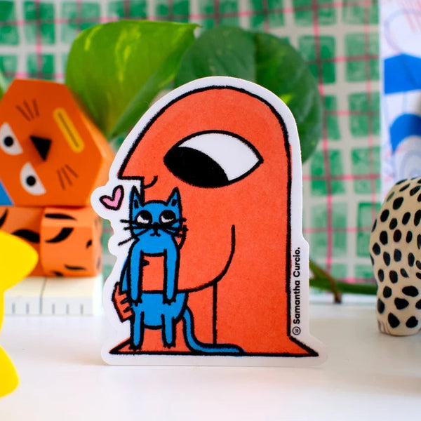 A die-cut sticker featuring an orange figure holding a blue cat, displayed on a tabletop with colourful toys and green leaves in the background.