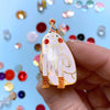 Hand holding a decorative pin with a cartoon-like character wearing a party hat against a colouful confetti background.