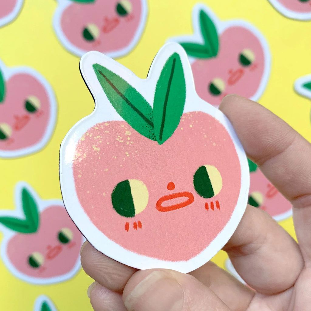 Hand holding a cute peach magnet with a yellow background featuring more similar magnets.