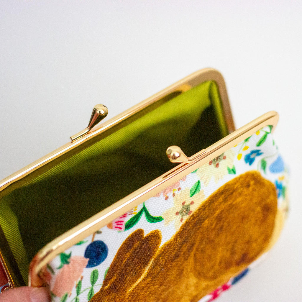 A close-up of an open kiss-lock clutch purse with a white fabric exterior featuring a colourful floral print and a large brown rabbit motif, a bright green cotton-lined interior, and a gold-tone metal frame.