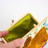 A close-up of a hand holding open a small clutch purse. The purse has a gold-tone metal frame, a bright olive green cotton interior lining, and a white floral exterior with a brown pattern on the flap.