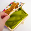 A close-up of a hand holding open a small clutch or coin purse with a gold-tone metal frame and a bright green cotton interior. The exterior fabric has a multicolour floral print with a large brown animal illustration.