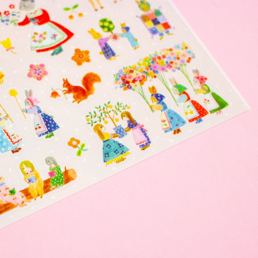 A close-up of a sheet of decorative stickers featuring whimsical illustrated characters, flowers, and animals in bright pastel and saturated colours on a white background, set against a solid pink backdrop.