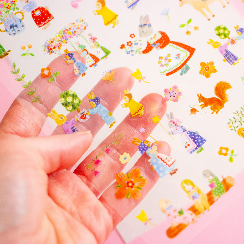 A hand holds a clear sheet of small illustrated stickers or decals featuring colourful woodland characters, flowers, and animals.