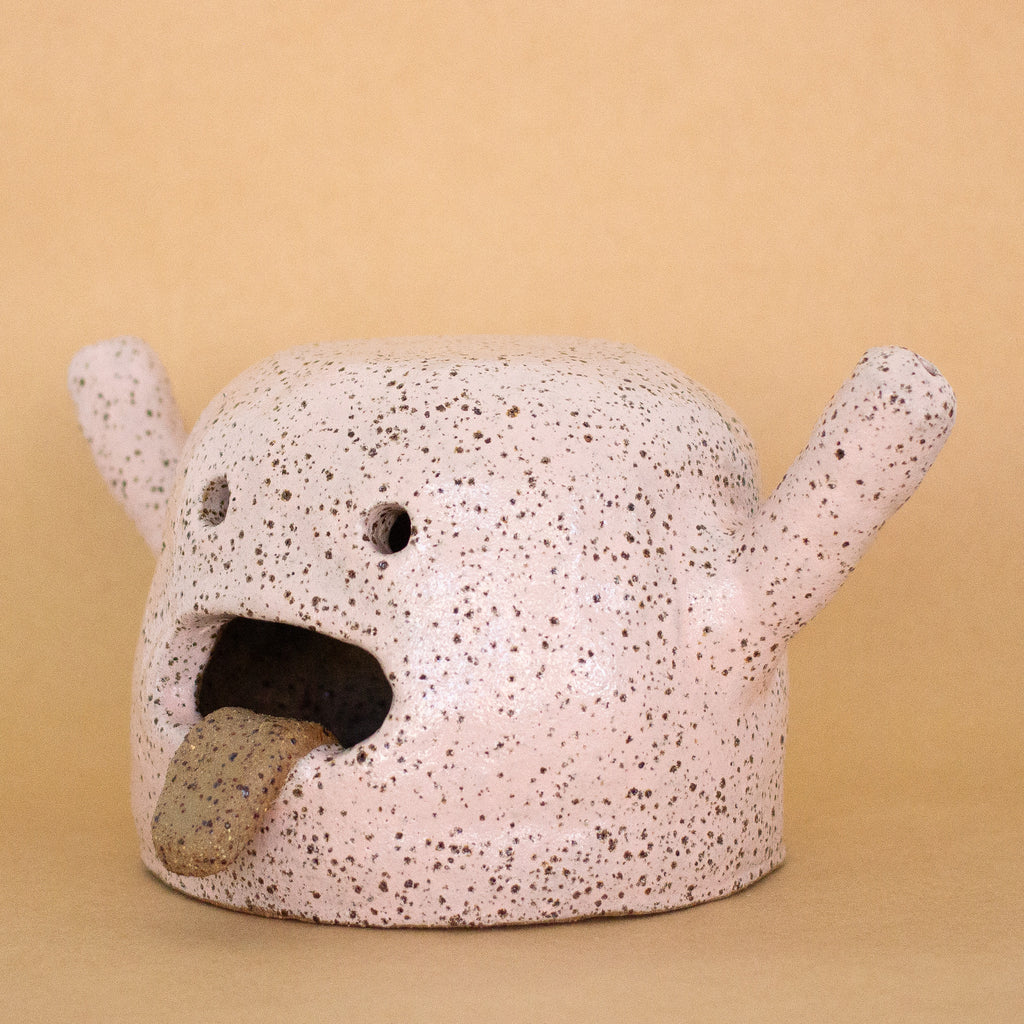 Side facing photo of the rah figurine. Artwork is made from stoneware clay with a pink glaze. Figurine has an open mouth with tongue hanging out, two eyes, and two hands that are extended. 