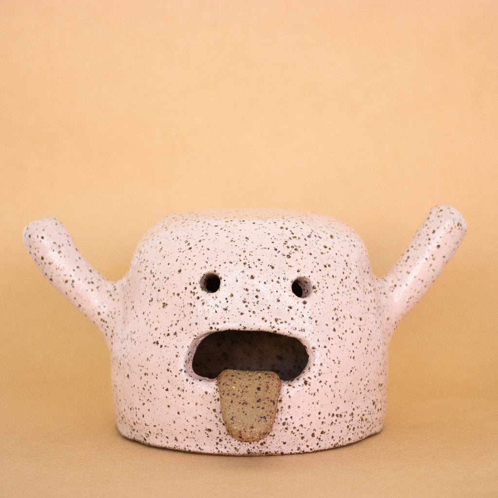 Front facing photo of the rah figurine. Artwork is made from stoneware clay with a pink glaze. Figurine has an open mouth with tongue hanging out, two eyes, and two hands that are extended. 