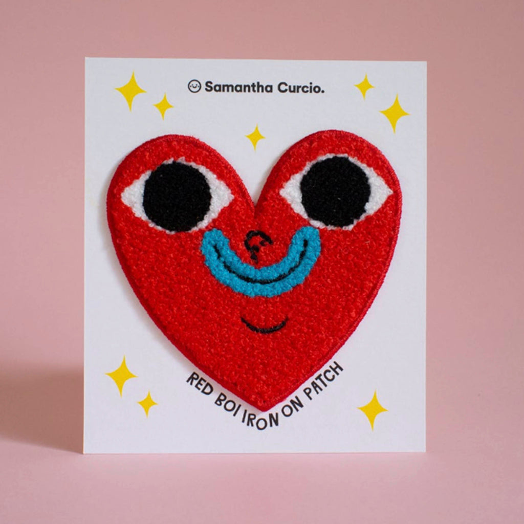 A red heart-shaped iron-on patch with embroidered eyes, a blue smile, and black curved lines, shown on a white backing card that reads 'Samantha Curcio.' and 'RED BOI IRON ON PATCH,' against a soft pink background.