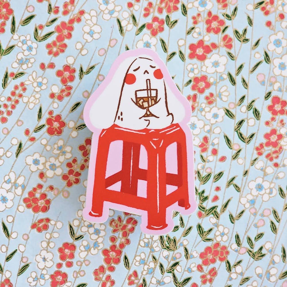 A die-cut sticker featuring a cartoon character with a white bunny holding an iced drink, sitting on a red stool, placed on a light blue floral background.