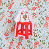 A die-cut sticker featuring a cartoon character with a white bunny holding an iced drink, sitting on a red stool, placed on a light blue floral background.