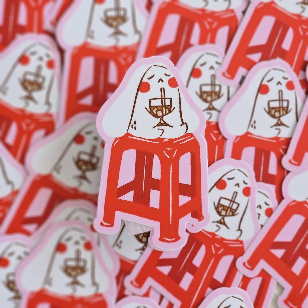 A close-up pile of die-cut stickers featuring a white bunny-like character with red cheeks holding a ice drink, seated on a red stool. The stickers have pink border, with multiple copies scattered in the background.