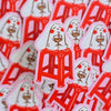 A close-up pile of die-cut stickers featuring a white bunny-like character with red cheeks holding a ice drink, seated on a red stool. The stickers have pink border, with multiple copies scattered in the background.
