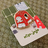A Misu Juju enamel pin shaped like a red plastic stool with a cute cartoon character inside, mounted on its illustrated backing card.