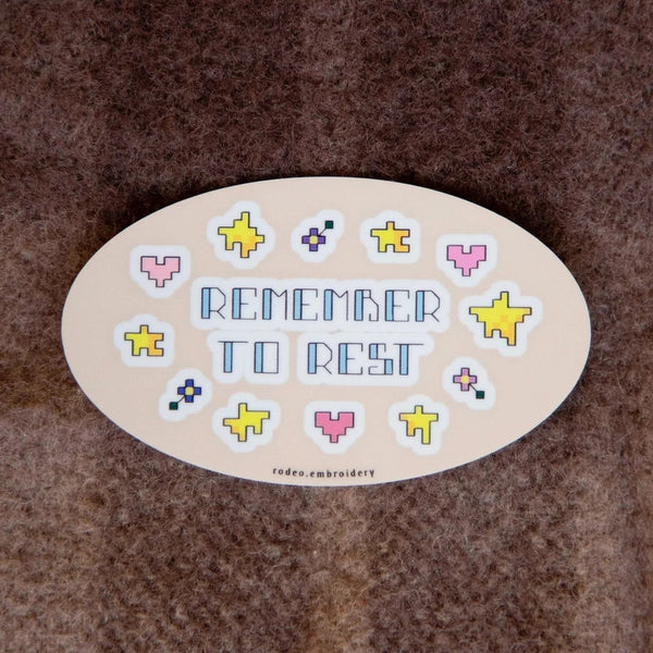 An oval cream-coloured embroidered patch with the words 'REMEMBER TO REST' in blue, surrounded by small pixel-art style hearts and stars in pink, yellow, and purple, placed on a brown textured fabric background.