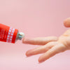 Hand applying cream from a red tube labeled 'Restore Hanami' on a pink background