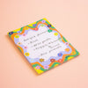 A rectangular notepad with a colourful floral border and a white lined center, showing a handwritten recipe. The notepad is photographed on a solid peach-pink background.