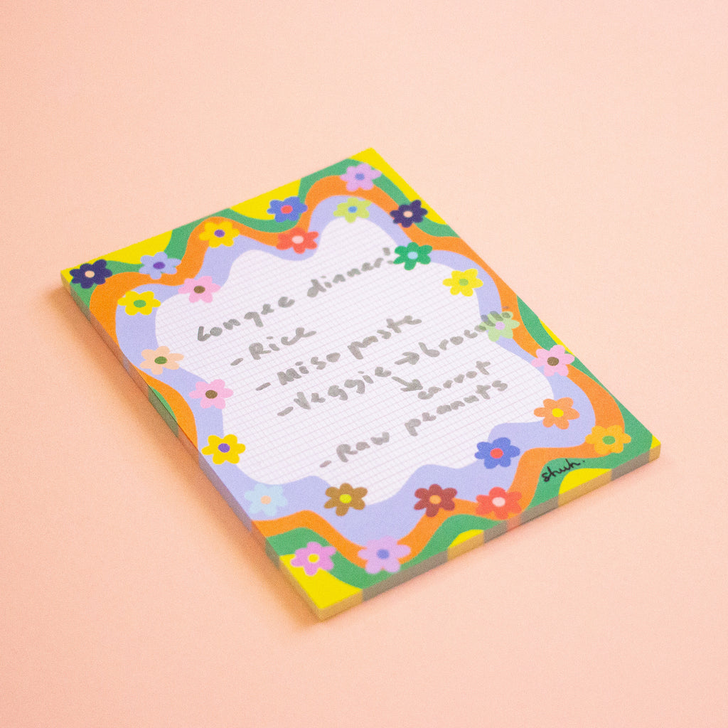 A rectangular notepad with a colourful floral border and a white lined center, showing a handwritten recipe. The notepad is photographed on a solid peach-pink background.