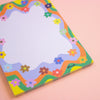 A rectangular notepad with a colourful retro wavy border and small flowers, photographed on a soft pink background. The notepad has a white grid pattern in the center and the brand name 'shuh.