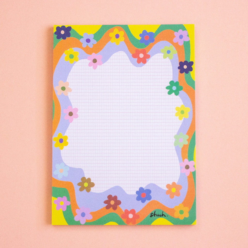 A rectangular notepad with a colourful retro floral border and a white grid writing area, photographed flat against a soft peach background.