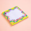 A square sticky note pad with a colourful retro floral border on a soft peach background. The pad features a white mesh writing area surrounded by wavy bands of purple, green, and orange, with a bright yellow outer border decorated with small flowers.