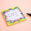 A square sticky note pad with a colourful floral border and the handwritten message 'You mean the world to me!' on a pale pink background, with a black pen partially visible at the right edge.
