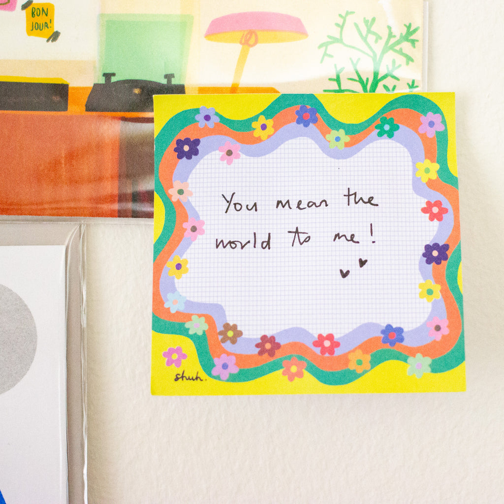 A square handmade paper note with a colourful floral border and handwritten text reading 'You mean the world to me!' pinned to a light-coloured wall.