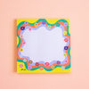 A square notepad with a colourful retro wavy border and small flowers, photographed flat against a soft peach background.