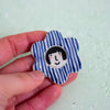 Blue and white striped flower shaped brooch with a face design held by a hand.