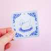 A hand holding a square white sticker with a blue hand-drawn swan in the center and a decorative blue floral border, set against a solid light pink background.
