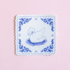 A square white coaster with scalloped edges, featuring a blue line drawing of a swan in a pond and a matching blue floral border, set against a solid light pink background.