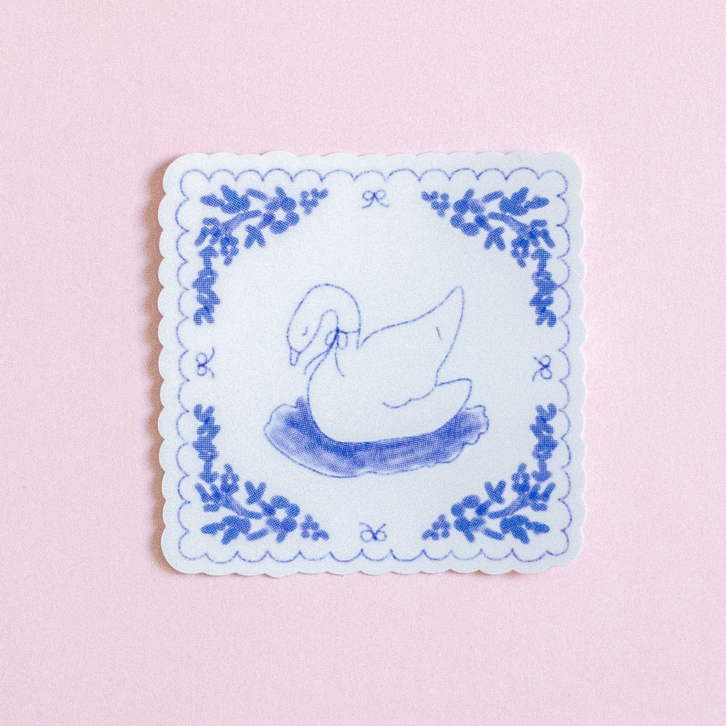 A square white coaster with scalloped edges, featuring a blue line drawing of a swan in a pond and a matching blue floral border, set against a solid light pink background.