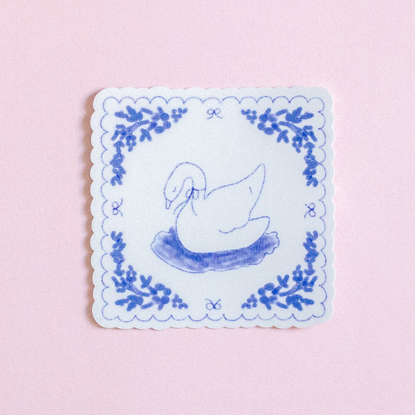 A square white coaster with scalloped edges, featuring a blue line drawing of a swan in a pond and a matching blue floral border, set against a solid light pink background.