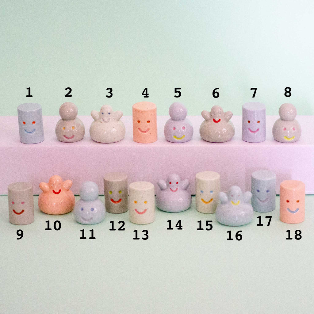 A set of pastel-coloured ceramic figurines and small cylinders with simple smiley faces, arranged in two rows on a pale mint background. Each piece is numbered from 1 to 18 with black text above and below.