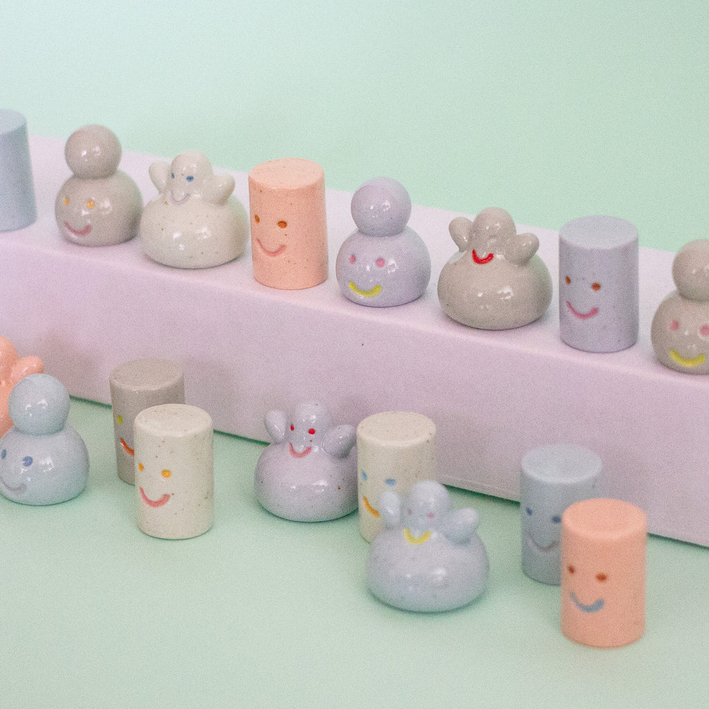A collection of pastel-coloured, speckled ceramic figurines with simple painted faces, arranged on a light mint green surface and a white display riser.