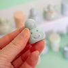 A hand holding a small, glossy, light blue smiling cloud-shaped trinket against a soft-focus pastel background.