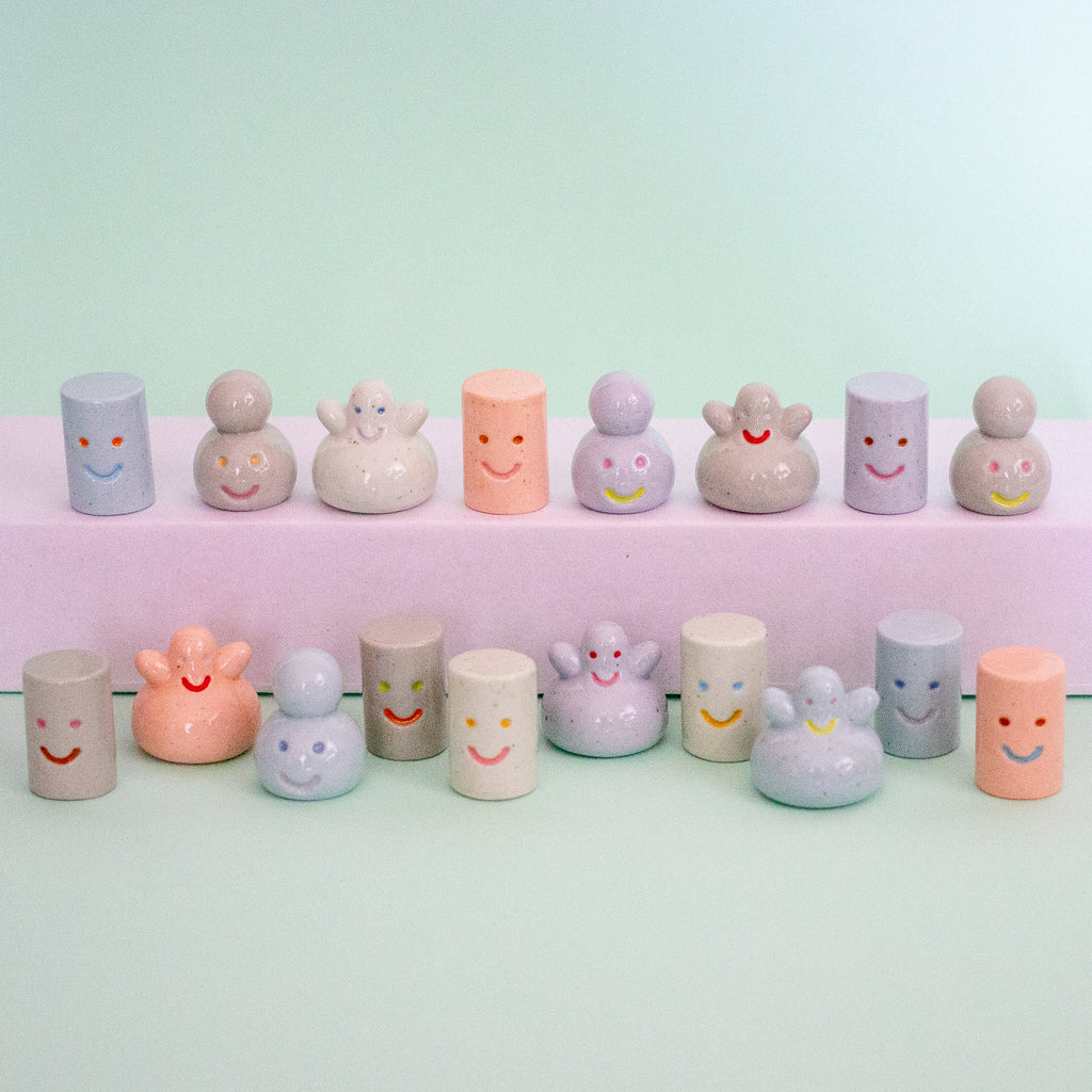 A set of pastel-coloured ceramic figurines with simple smiley faces, arranged on a light mint green background with a white display block behind them.