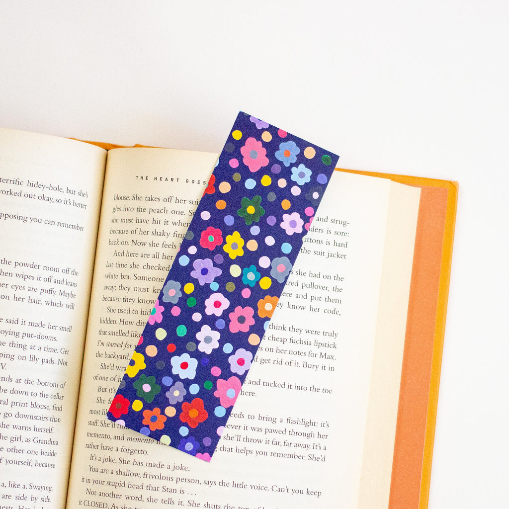 A floral book bookmark with a dark blue background and colourful polka-dot flowers, shown draped over an open book on a white background.