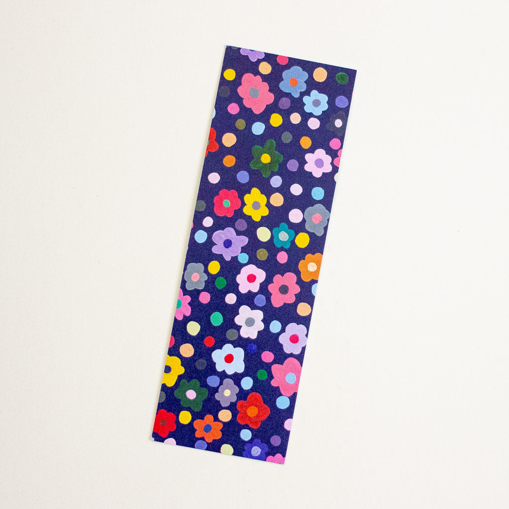 A rectangular bookmark, featuring a colourful floral and polka dot pattern on a dark navy background, shown on a plain off-white surface.