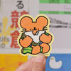 A hand holding a sticker of a mouse cartoon eating a sandwich against a colourful background. 