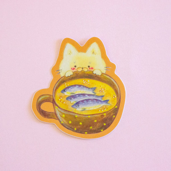 A die-cut sticker featuring a cute illustration of a cream-coloured cat peeking over a bowl of soup with fish on top, set against a solid light pink background.