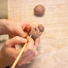 Close-up of a person's hands using a wooden sculpting tool to refine a small clay animal figurine, with clay balls blurred in the background on a light work surface.