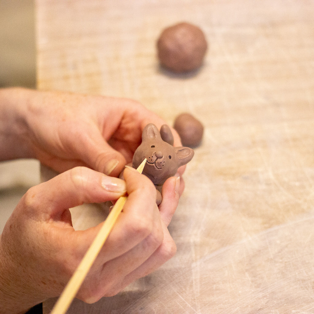 Close-up of a person's hands using a wooden sculpting tool to refine a small clay animal figurine, with clay balls blurred in the background on a light work surface.
