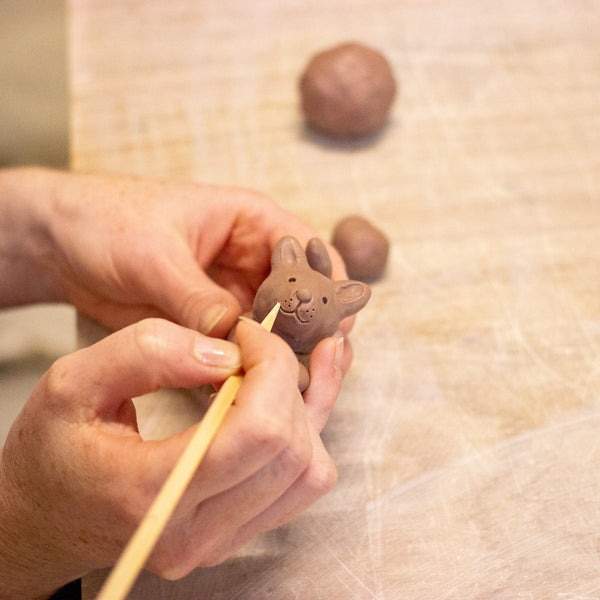 Close-up of a person's hands using a wooden sculpting tool to refine a small clay animal figurine, with clay balls blurred in the background on a light work surface.