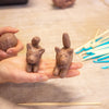 A close-up of a person's hands holding two small clay animal figurines shaped like cats, with pottery tools on a wooden surface in the background.