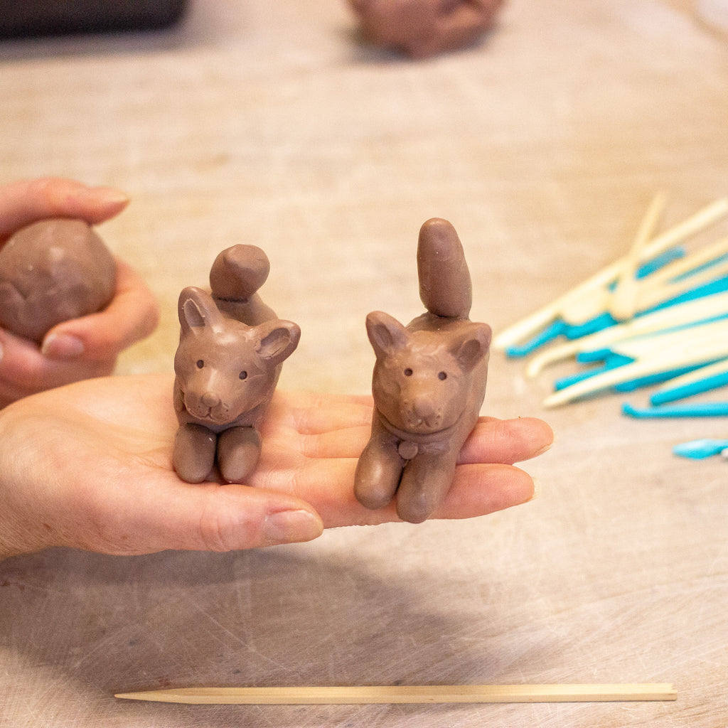 A close-up of a person's hands holding two small clay animal figurines shaped like cats, with pottery tools on a wooden surface in the background.
