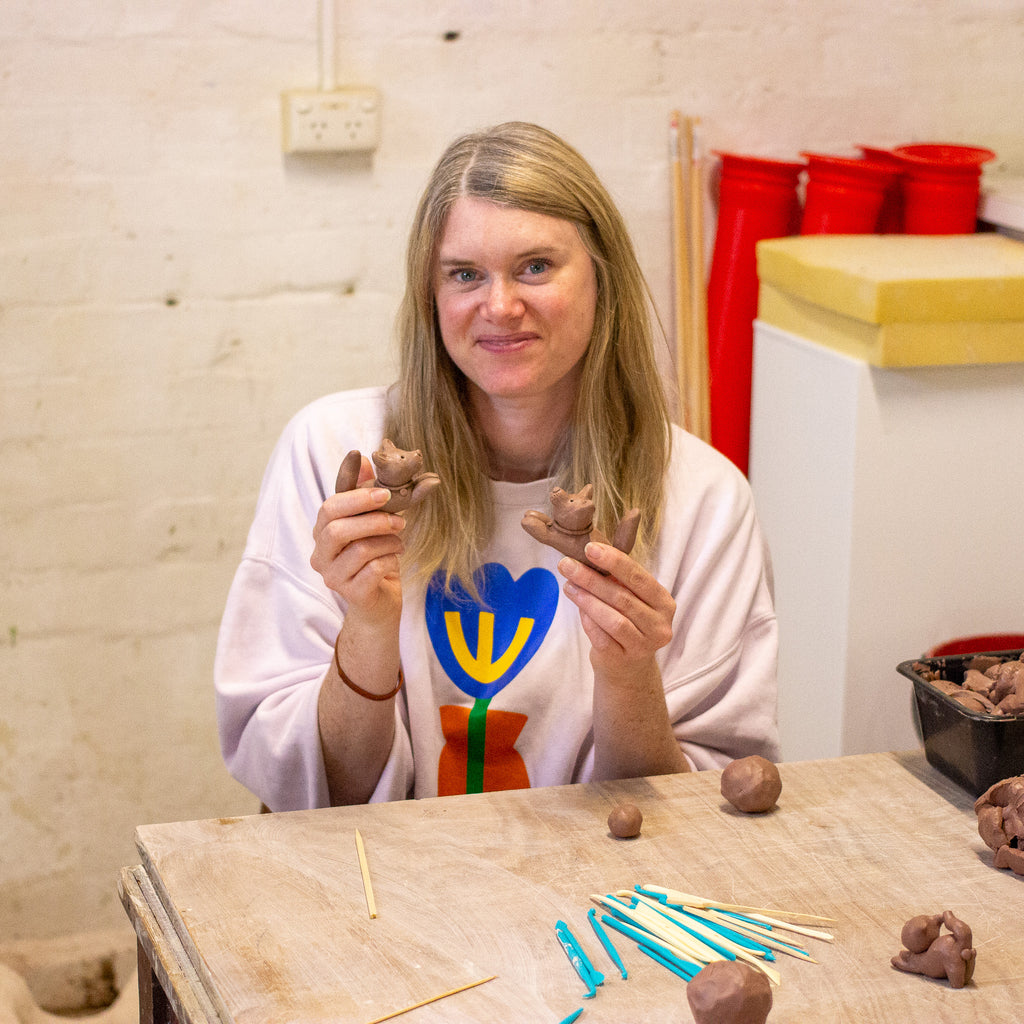 A woman with blonde hair sits at a wooden table in a pottery studio, holding two small clay animal figures. She wears a white sweatshirt with a colourful tulip graphic.