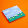 A square sticky note featuring a painted landscape of a blue sky with clouds and a green field of colourful wildflowers, displayed on a solid orange background.